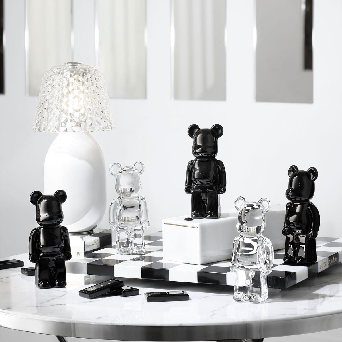 Five Bear Brick glass figurines by Baccarat, in black and clear, are displayed on a glossy white table amid geometric decor, a small lamp, mirrors, and rectangular accents—a refined showcase of luxury collectible bear art toys.