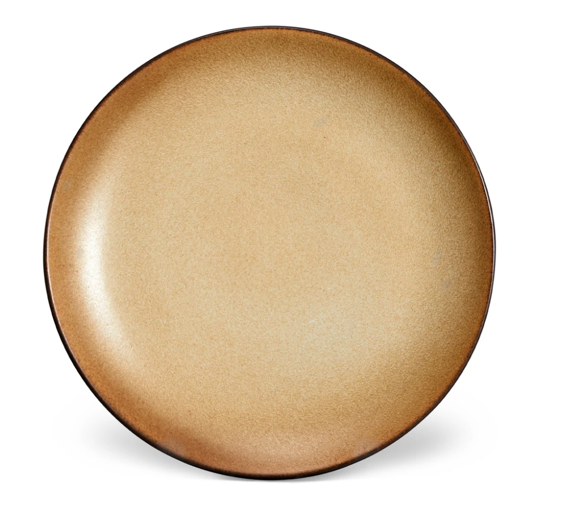 The Terra Leather Dinnerware by L'Objet features a smooth, light brown ceramic plate with a darker rim. Its elegant glazed finish and round design make it ideal for displaying hand-crafted table settings.