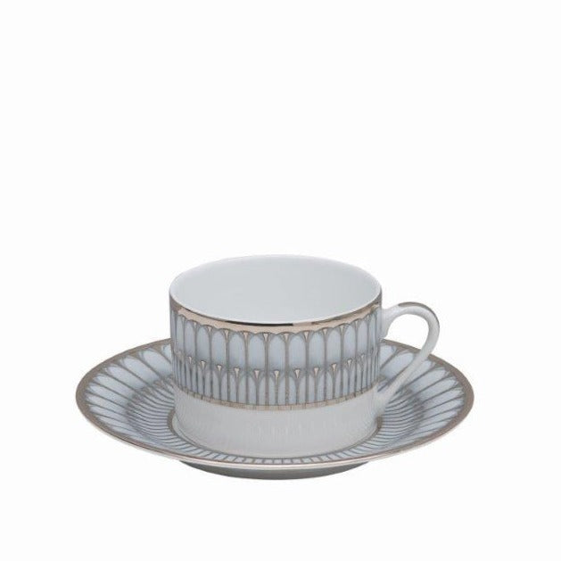 A white Philippe Deshoulieres BIA Arcades Platinum teacup with handle sits on a matching saucer, both crafted from fine Limoges porcelain and featuring an elegant gold and gray geometric rim design. The background is plain white.
