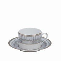 A white Philippe Deshoulieres BIA Arcades Platinum teacup with handle sits on a matching saucer, both crafted from fine Limoges porcelain and featuring an elegant gold and gray geometric rim design. The background is plain white.