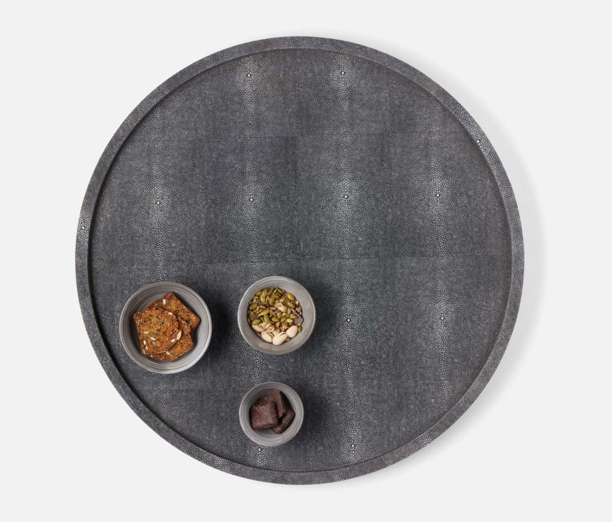 The Calvin Faux Shagreen Lazy Susan in grey by Blue Pheasant holds three small bowls with crackers, mixed nuts, and dark chocolate, all arranged on a white background.