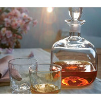 A Simon Pearce Woodbury Bourbon decanter sits on a table next to two glasses—one partly filled with amber liquor. Sunlight filters in, with blurred pink flowers in the background.