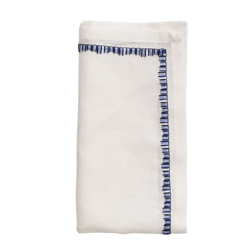 The Kim Seybert Filament Napkin White & Navy Set of 4 features neatly folded white linen napkins with blue embroidered borders, shown against a plain white background.
