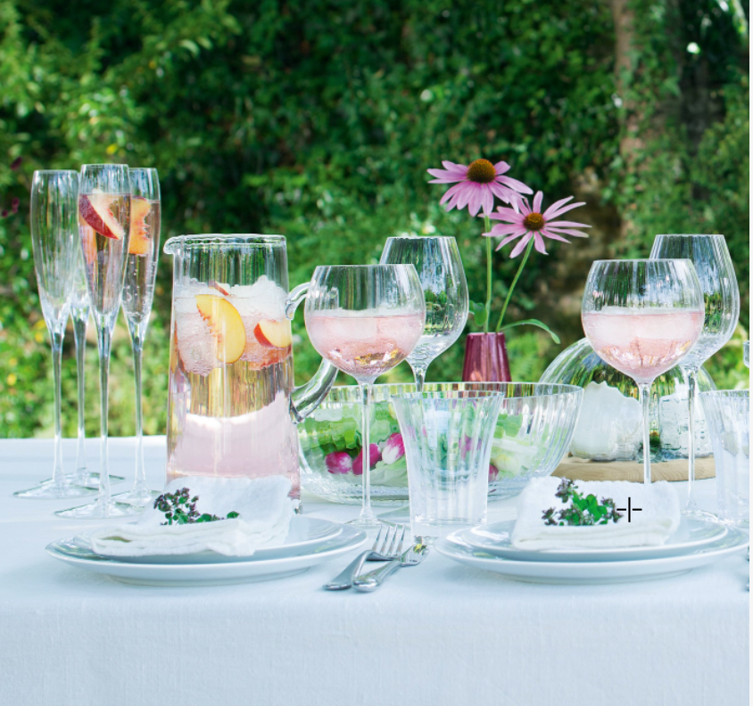 A white cloth-covered outdoor table features LSA’s Aurelia Jug filled with fruit slices, glasses of pink drink, plates with napkins and herbs, mouth-blown glass bowls, a vase of purple flowers, and a lush green foliage backdrop.