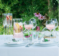 A white cloth-covered outdoor table features LSA’s Aurelia Jug filled with fruit slices, glasses of pink drink, plates with napkins and herbs, mouth-blown glass bowls, a vase of purple flowers, and a lush green foliage backdrop.