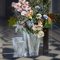 The Baccarat Eye Rectangular Vase Clear, featuring a textured design, holds a colorful bouquet and stands before a mirror, reflecting its beauty on a geometric-patterned floor.
