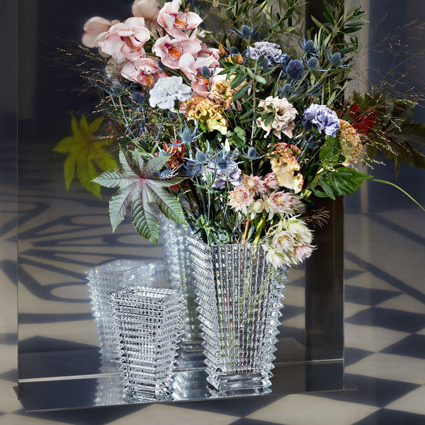 The Baccarat Eye Rectangular Vase Clear, featuring a textured design, holds a colorful bouquet and stands before a mirror, reflecting its beauty on a geometric-patterned floor.
