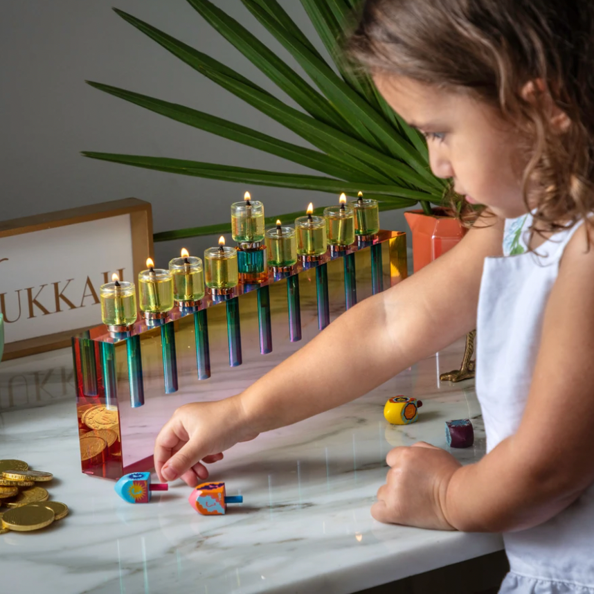 A young girl spins colorful dreidels on a marble table beside the Apeloig Modern Acrylic Oil Menorah with lit candles, chocolate coins, and a festive sign. A green plant and flower vase complete the cheerful Hanukkah scene in the background.