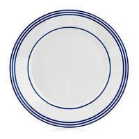 The Latitudes Dinnerware by Royal Limoges is a handmade white ceramic plate featuring three blue concentric rings around the rim, shown from above on a white background.