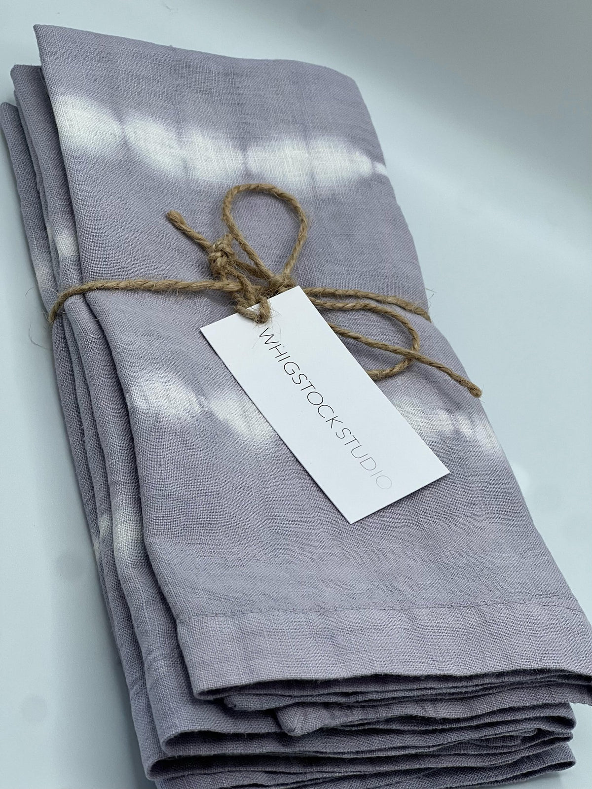A set of four hand dyed linen napkins in lavender gray by Whigstock Studio is neatly folded, tied with brown twine, and features a branded tag, displayed against a light background.
