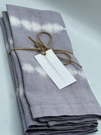 A set of four hand dyed linen napkins in lavender gray by Whigstock Studio is neatly folded, tied with brown twine, and features a branded tag, displayed against a light background.