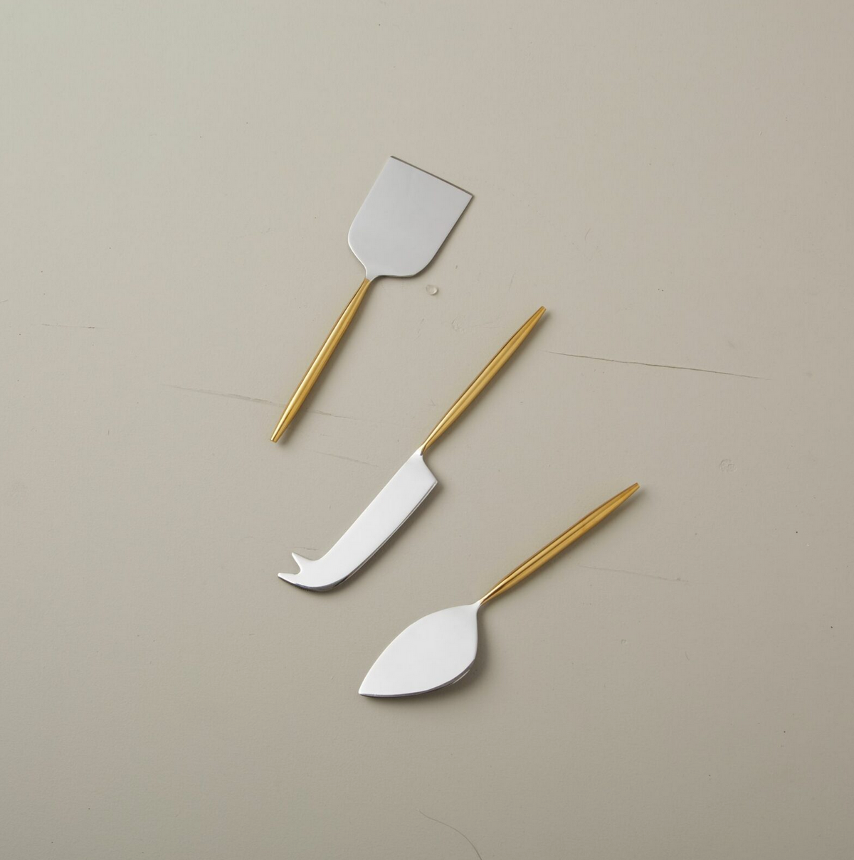 The Be Home Mixed Metal Cheese Set – Silver & Gold features three cheese knives with gold handles and silver blades, each designed with a unique shape for serving various types of cheese.