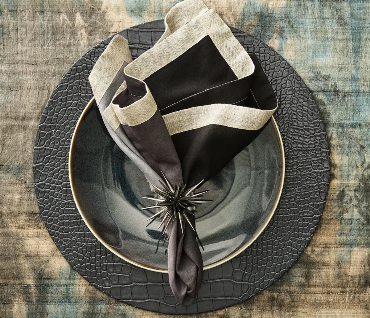 A dark gray plate rests on the Kim Seybert Croco Placemat Charcoal (Set of 4), paired with a two-tone napkin and a spiky napkin ring, all arranged on a rustic wooden table.