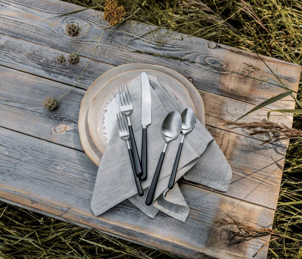 A place setting with a grey napkin, two plates, and the Simon Pearce Hampton Flatware 5 Piece Setting rests on a rustic wooden table outdoors.