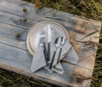 A place setting with a grey napkin, two plates, and the Simon Pearce Hampton Flatware 5 Piece Setting rests on a rustic wooden table outdoors.