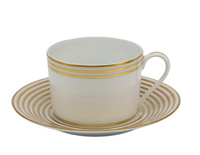 A white Latitudes teacup by Royal Limoges features gold horizontal stripes near the rim and sits on a matching saucer adorned with concentric gold rings on a white background.