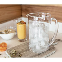 A Simon Pearce Ascutney Pitcher Medium filled with ice cubes sits on a wooden cutting board. Nearby are a glass of orange smoothie, a bowl of yogurt with seeds, a spoon, and halved oranges on a light kitchen counter.