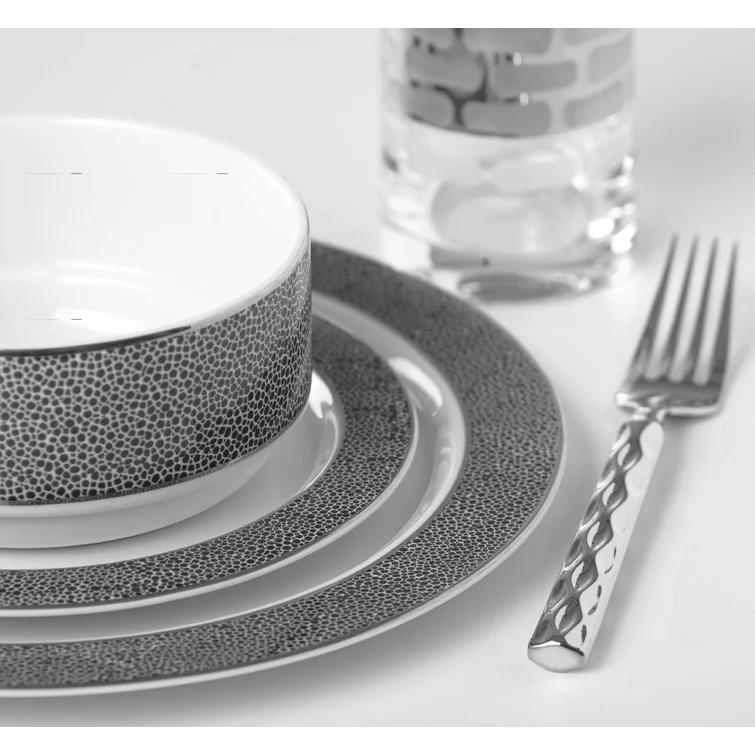 Three textured, patterned plates and a matching bowl from the Michael Wainwright Panthera Dinnerware Platinum collection are stacked with a modern glass and a textured-handle fork, all arranged on a white surface.