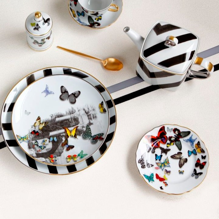 The Sol Y Sombra tea set by Christian Lacroix x Vista Alegre features black and white stripes, colorful butterflies, a large charger, small plate, teapot, sugar bowl, and gold spoon on a light textured surface.
