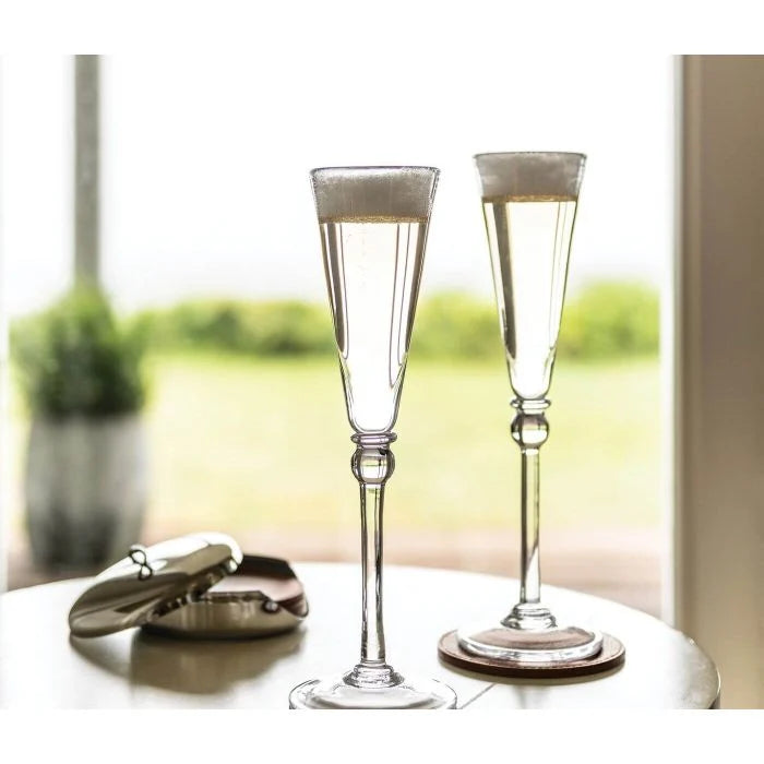 Two Simon Pearce Hartland Flute glasses filled with bubbly sit on a round table by a compact mirror, greenery, and sunlight—an elegant handmade glassware set of two, perfect for gifting at weddings.