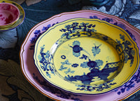 A close-up of the Oriente Italiano Dinner Plate Azalea by Ginori 1735, featuring intricate blue floral and geometric designs on yellow with a pink rim, layered over a matching plate, with part of a small pink dish visible above.