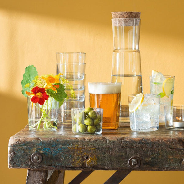 A rustic wooden table displays the LSA Gio Tumbler Large Set of 4, a carafe of water, a glass of beer, green olives, two lemon drinks on ice, flowers in a vase, and a lit candle, all set against a yellow wall.