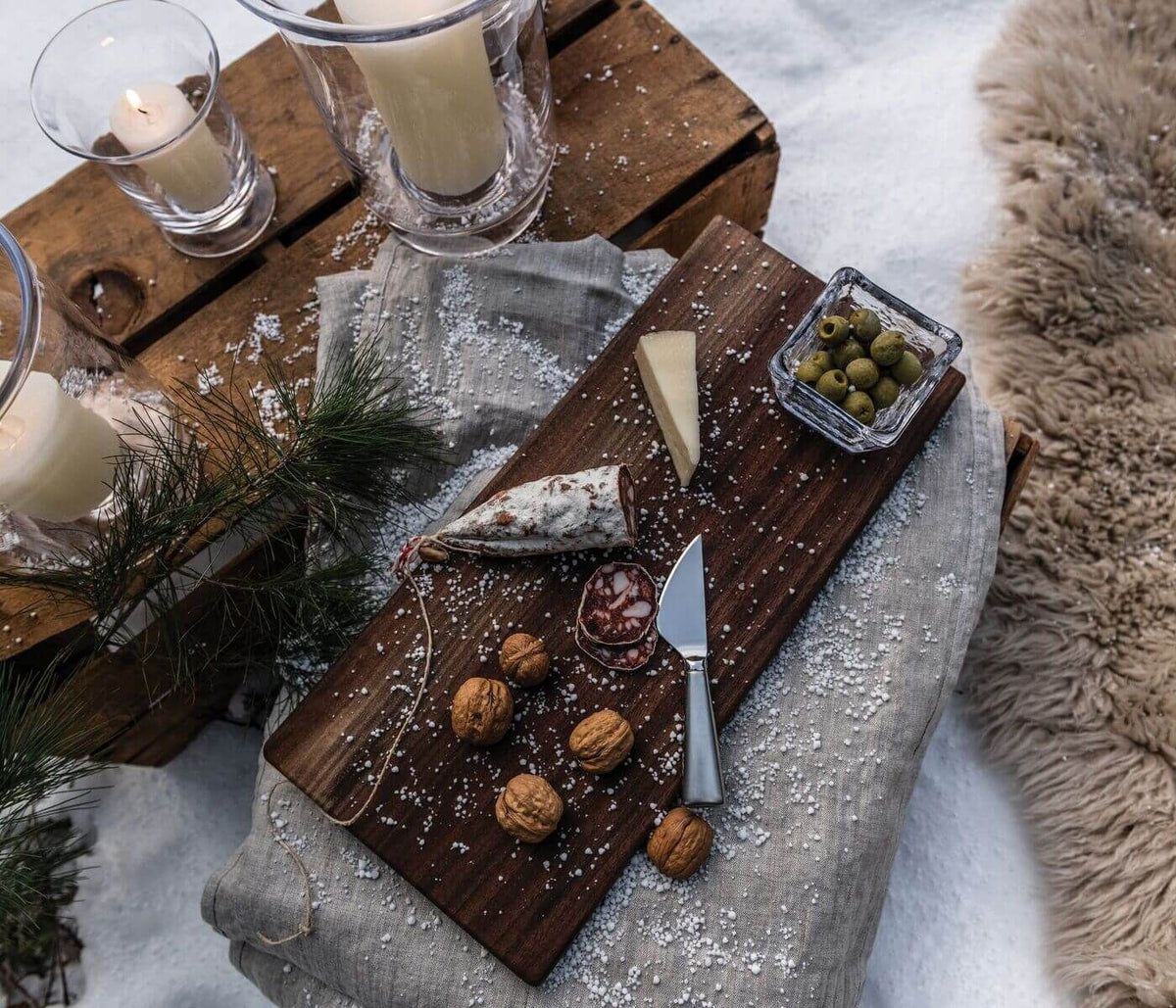 A rustic wooden board on snow holds walnuts, cheese, salami, green olives, dip, and the Simon Pearce Hartland Cheese Knife Set. Candles, pine branches, and a fur rug create a cozy winter outdoor scene.
