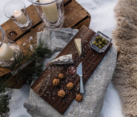 A rustic wooden board on snow holds walnuts, cheese, salami, green olives, dip, and the Simon Pearce Hartland Cheese Knife Set. Candles, pine branches, and a fur rug create a cozy winter outdoor scene.