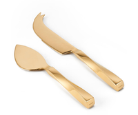 The Leon Gold Cheese Set by Aerin features two gold-colored cheese knives: a brass-plated stainless steel serrated knife with a forked tip and a matching flat-bladed server, both with angular handles and contemporary design.