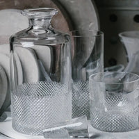 The Richard Brendon Diamond Decanter Large rests beside two matching glasses and a small, broken glass, all arranged with stacked plates—perfect for whisky lovers and admirers of luxury crystal barware.