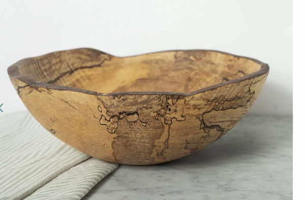 The Peterman Spalted Maple Oval Wood Bowl 18" features natural dark grain patterns and sits on a marble surface beside a folded striped cloth, set against a light background.
