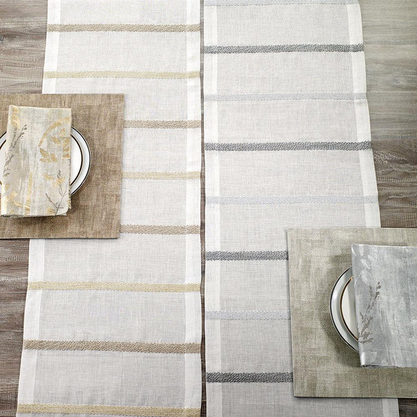 A top-down view of two table settings with Bodrum Metallic Band Silver Table Runners, each featuring a plate, a subtly patterned brown or gray napkin, and a matching rectangular placemat on a wooden table.