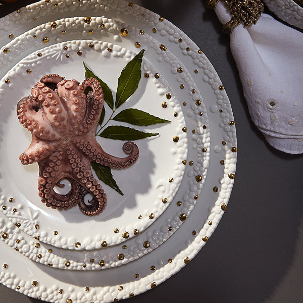 A cooked octopus sits on a decorative white L'Objet Haas Mojave Dinnerware Gold plate with gold accents and green leaves, stacked on matching fine porcelain plates. A nearby white napkin features a gold napkin ring.