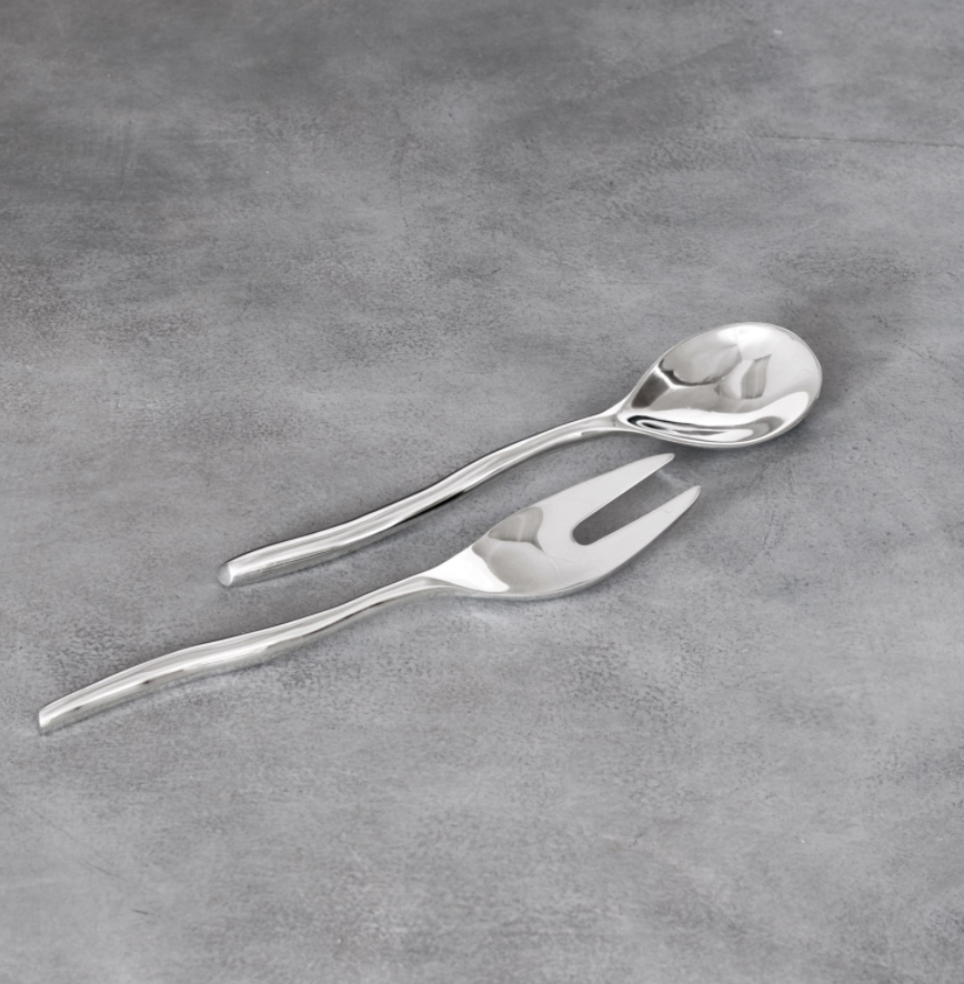 The Beatriz Ball Vento Claire Salad Set, featuring an artisan-crafted metal serving spoon and matching fork with gently curved handles, is displayed on a smooth gray surface.