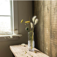 The Echo Lake Gather Vase by Simon Pearce, filled with yellow wildflowers and dried grasses, rests on a rustic wooden table near a window. Sunlight streams in, highlighting the artisan craftsmanship of this handmade glass vase.