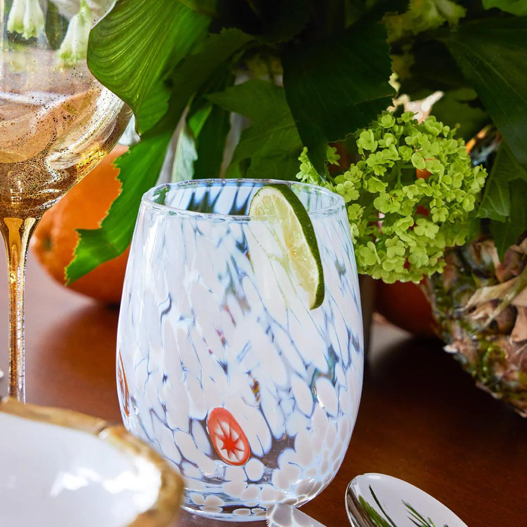 A close-up of a patterned Kim Seybert Gala Stemless Tumbler Set 4 with a lime wedge on the rim, on a wooden table featuring gold-rimmed glassware, a floral arrangement, and lush green leaves in the background.