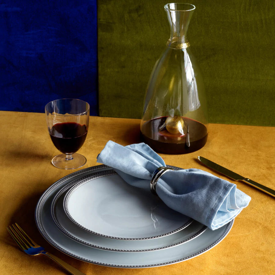 A table set with stacked white plates, L'Objet Linen Sateen Napkin Light Blue Set of 4 in a silver ring, red wine in a glass, a glass decanter, gold and silver cutlery on a mustard yellow tablecloth with blue and olive backgrounds.