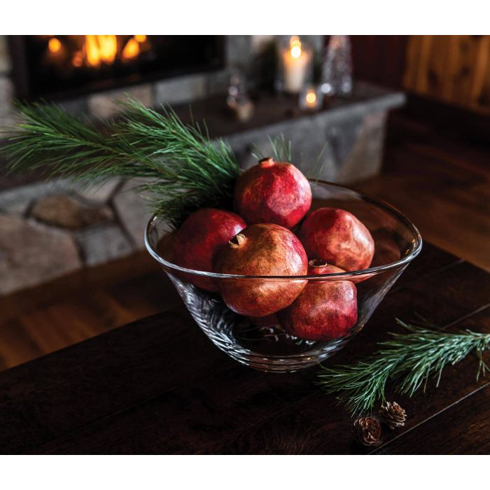 The Simon Pearce Barre Bowl, filled with pomegranates and pine branches, rests on a dark wooden table by the fireplace, surrounded by candles and stone decor for a cozy, rustic vibe.