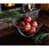 The Simon Pearce Barre Bowl, filled with pomegranates and pine branches, rests on a dark wooden table by the fireplace, surrounded by candles and stone decor for a cozy, rustic vibe.