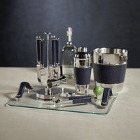 The Zodax Laguna Tool Bar Set features nickel bar accessories with navy blue accents, including a shaker, ice bucket, and utensils on a glass tray with a lime, displayed on a beige surface against a dark backdrop.