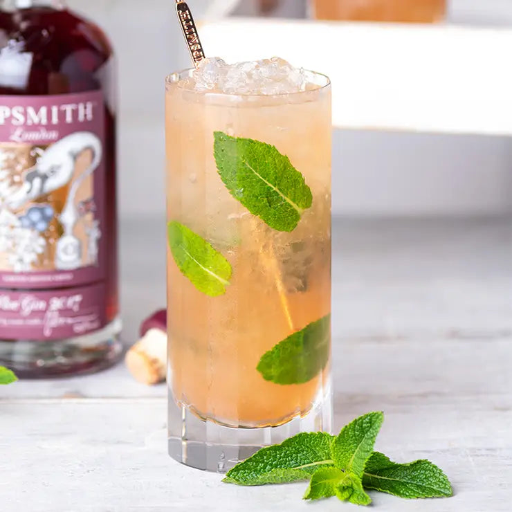 A Richard Brendon Fluted Highball filled with a light brown cocktail, ice, and mint sits on a white surface. A mint sprig is beside it, with barware and a Sipsmith gin bottle in the background.