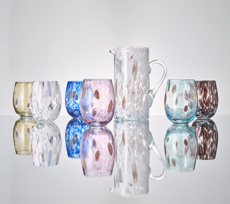 The Kim Seybert Gala Pitcher White and six colorful glass tumblers, crafted in Murano Glass style, are displayed on a reflective surface, creating mirrored reflections below them.