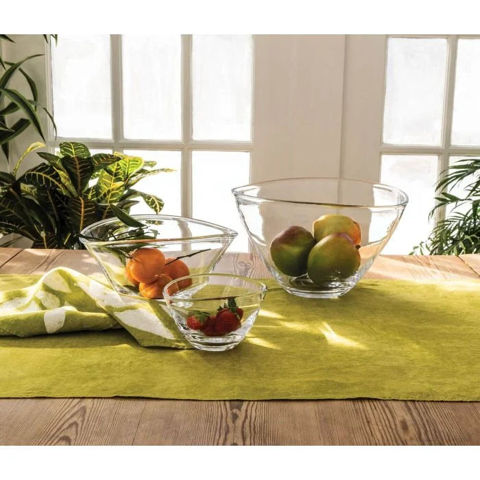 Three Simon Pearce Barre Bowls filled with mangoes, oranges, and strawberries are displayed on a green tablecloth near a sunlit window with potted plants in the background.