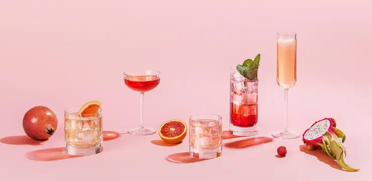 Five assorted cocktails, including one in the Richard Brendon Fluted Highball glass, are displayed on a pink surface amid pomegranate, blood orange halves, dragon fruit, and raspberries—highlighting elegant barware.