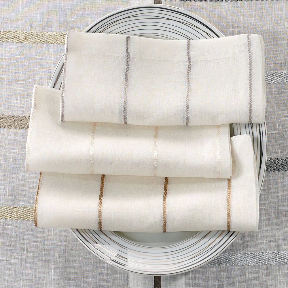 Four Bodrum Metallic Thread Napkin Pearl napkins are neatly arranged on a round white plate with gray circular lines, set atop a coordinating striped tablecloth.