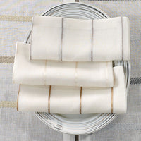 Four Bodrum Metallic Thread Napkin Pearl napkins are neatly arranged on a round white plate with gray circular lines, set atop a coordinating striped tablecloth.