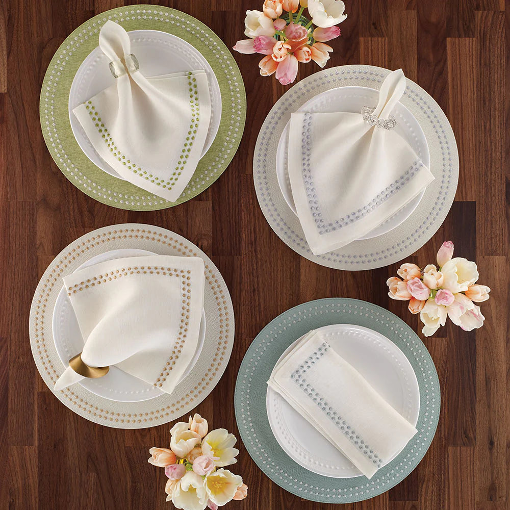 Four elegant place settings on a wooden table feature Bodrum Pearls Napkin Navy Set of 4, each paired with white plates with pastel accents, napkin rings, and a nearby bouquet of pink and white flowers.