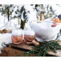Two glasses of sparkling rosé in Simon Pearce glassware rest on a wooden outdoor table, with a bottle of rosé chilling in the Simon Pearce Barre Bowl filled with snow. Pine branches, corks, and snowy trees set a wintry mood.