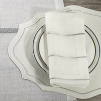 A close-up of a place setting featuring Bodrum’s Metallic Thread Napkin Pearl Set of 4, paired with a white scalloped placemat, a white plate with a gold rim, all on a gray and white textured tablecloth.