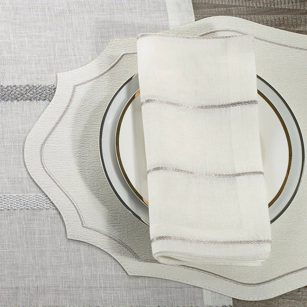 A close-up of a place setting featuring Bodrum’s Metallic Thread Napkin Pearl Set of 4, paired with a white scalloped placemat, a white plate with a gold rim, all on a gray and white textured tablecloth.